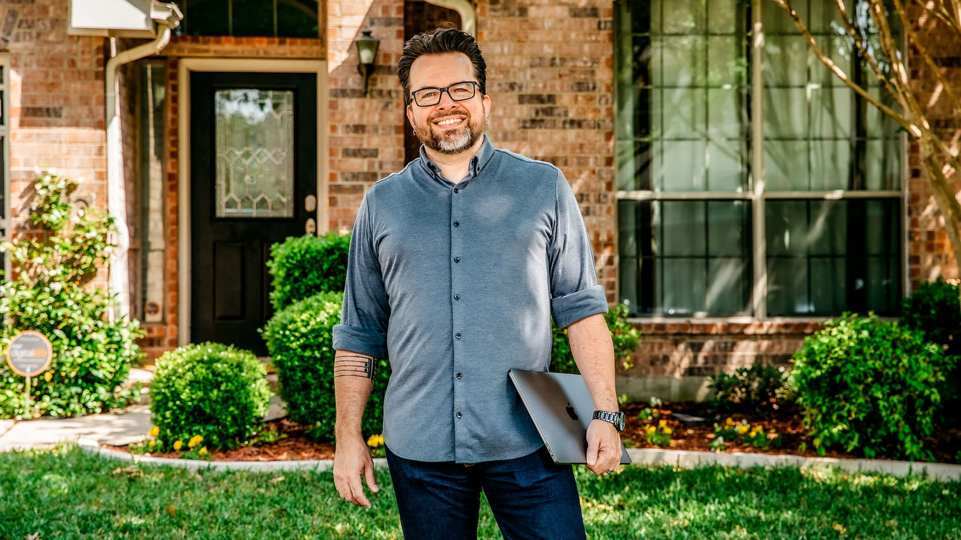 Boots standing in the front yard of his home, holding his laptop.